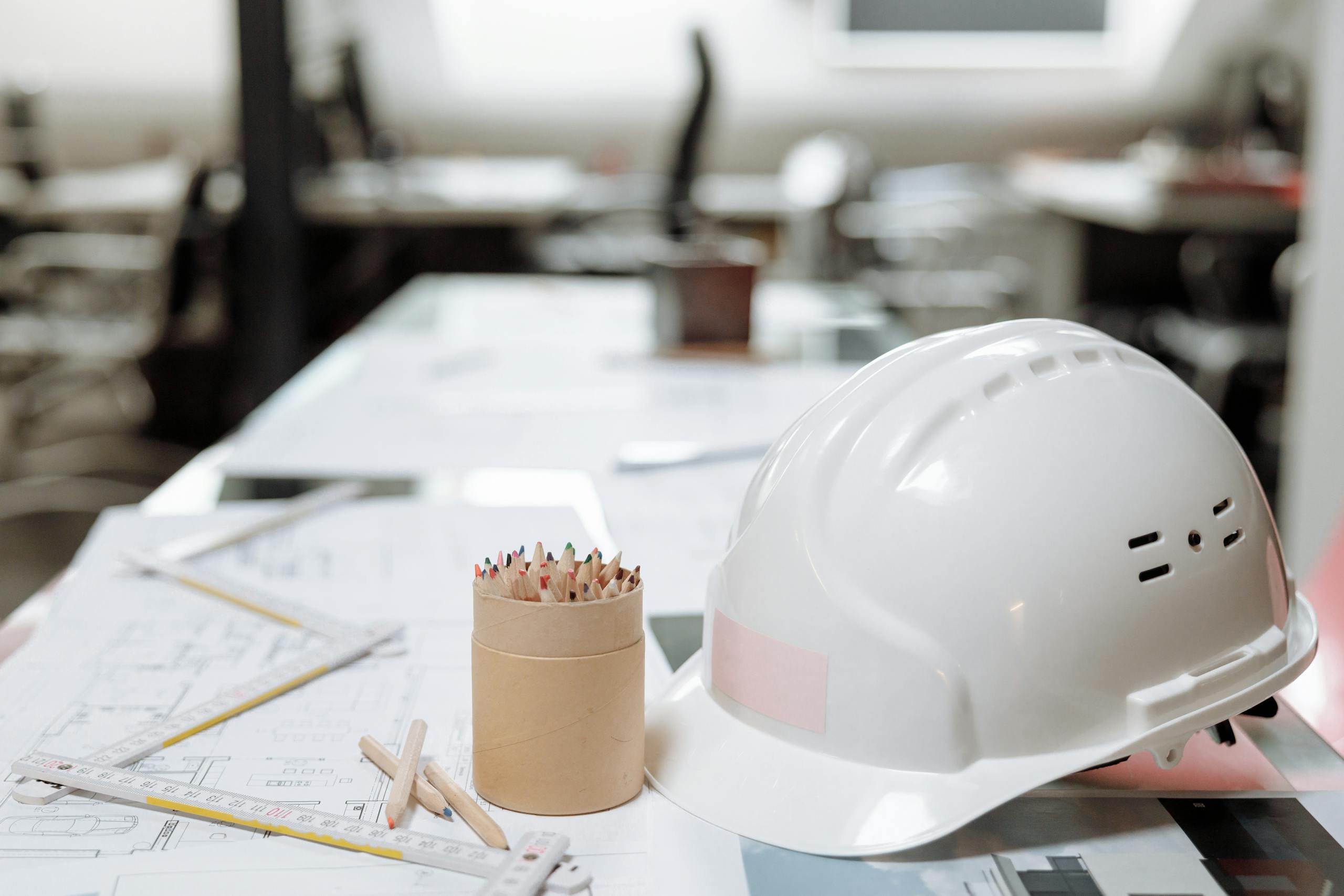 Close-up of an office desk with a white helmet, colored pencils, and architectural plans.